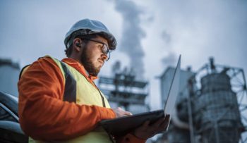 A shot of an young engineer wearing a helmet and using a laptop and hands free device during his night shirt in the oil rafinery. Engineering concept.